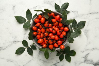Photo of Ripe rose hip berries with green leaves on white marble table, flat lay