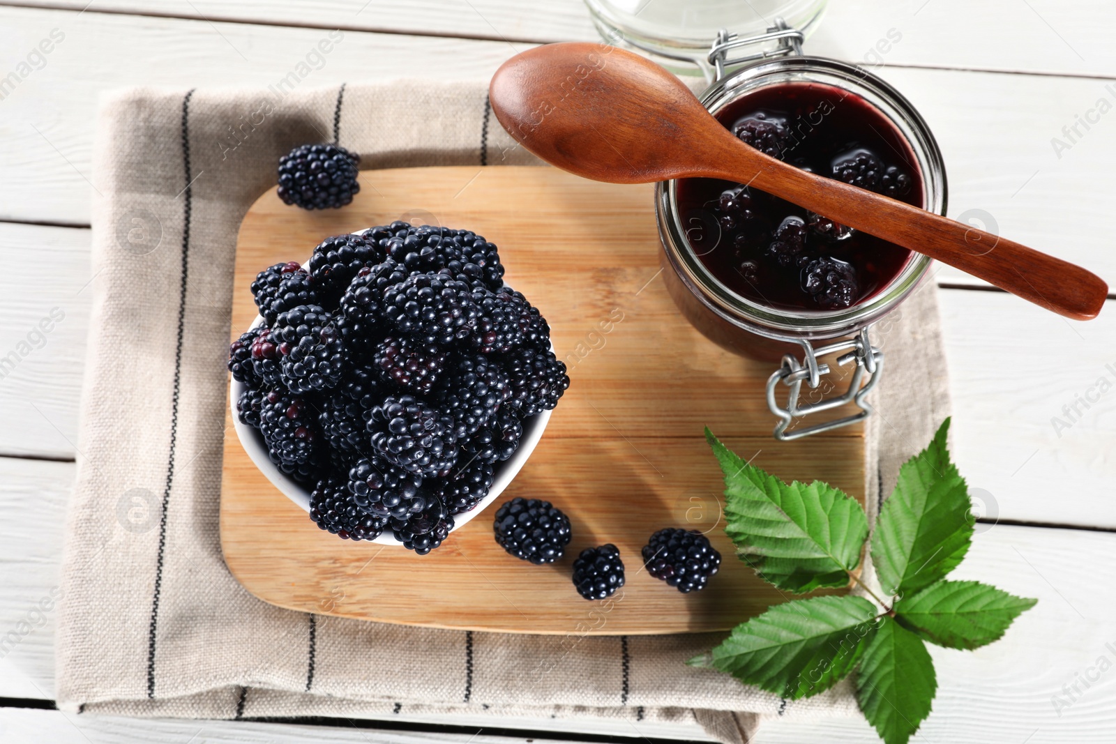 Photo of Fresh ripe blackberries, tasty jam and leaves on white wooden table, above view