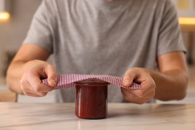 Man packing jar of jam into beeswax food wrap at light table indoors, closeup