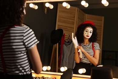 Photo of Young woman in mime costume putting gloves near mirror indoors
