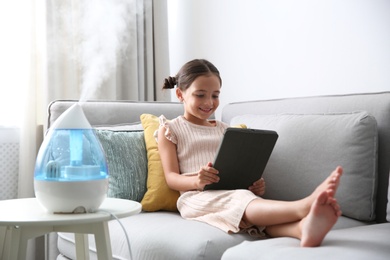 Photo of Little girl using tablet in room with modern air humidifier
