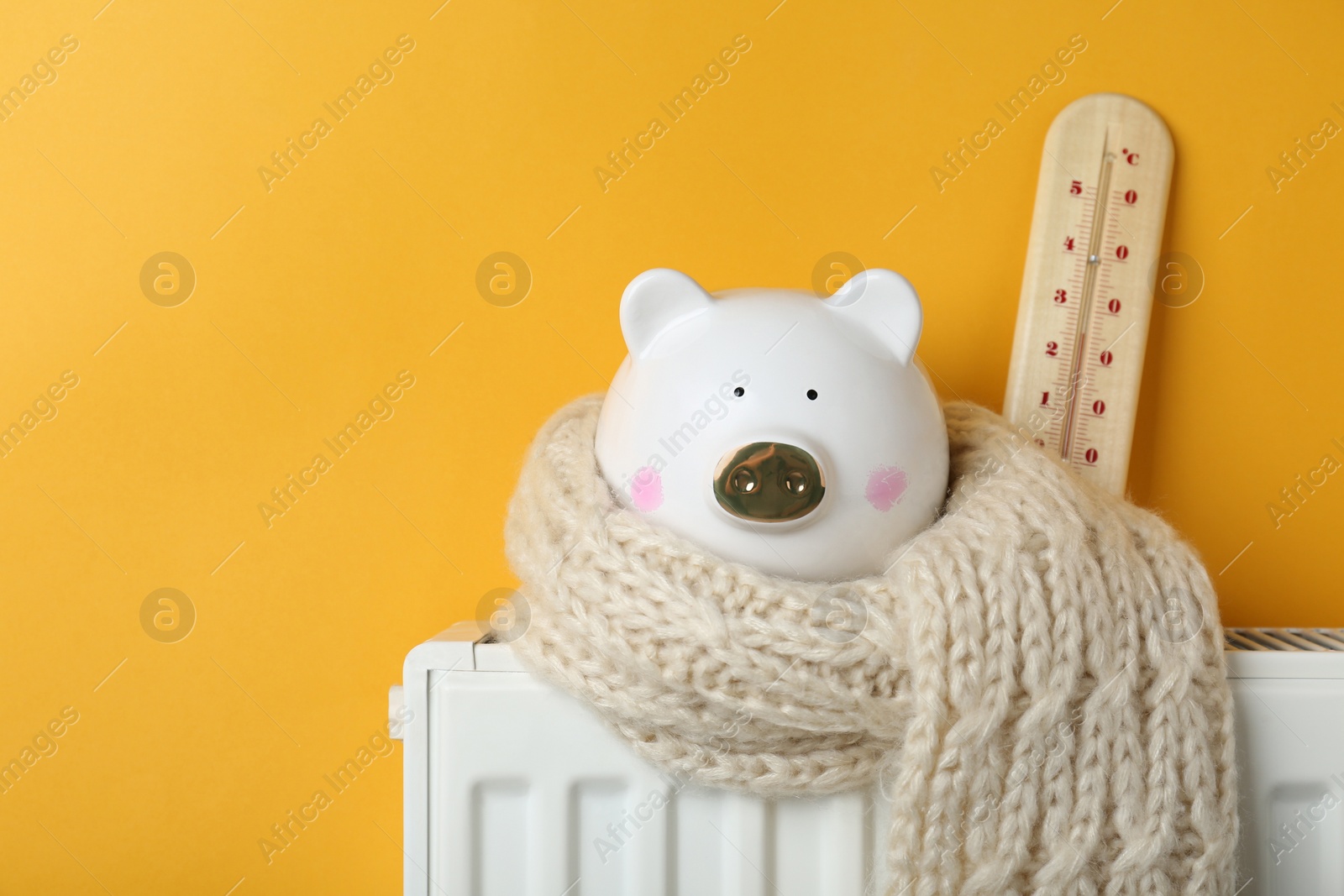 Photo of Piggy bank wrapped in scarf and thermometer on heating radiator against orange background