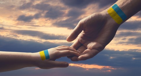 Image of Help for Ukraine. Man and child holding hands with drawings of Ukrainian flag and beautiful sky with clouds at sunset on background, closeup. Banner design