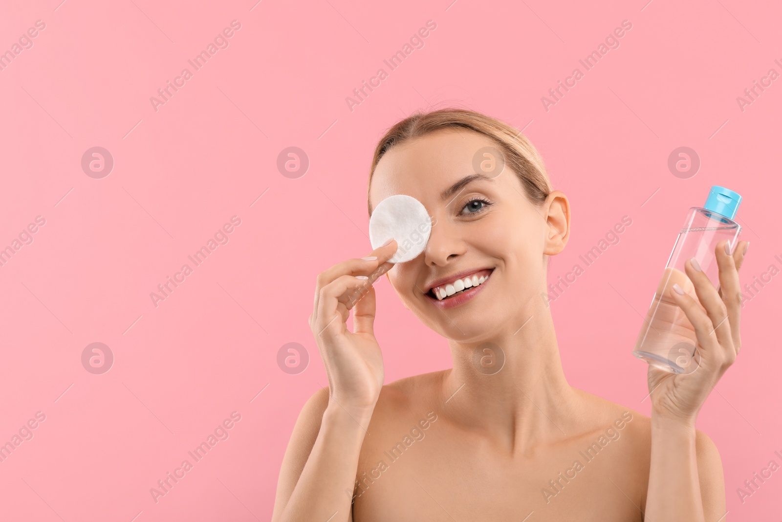 Photo of Smiling woman removing makeup with cotton pad and holding bottle on pink background. Space for text