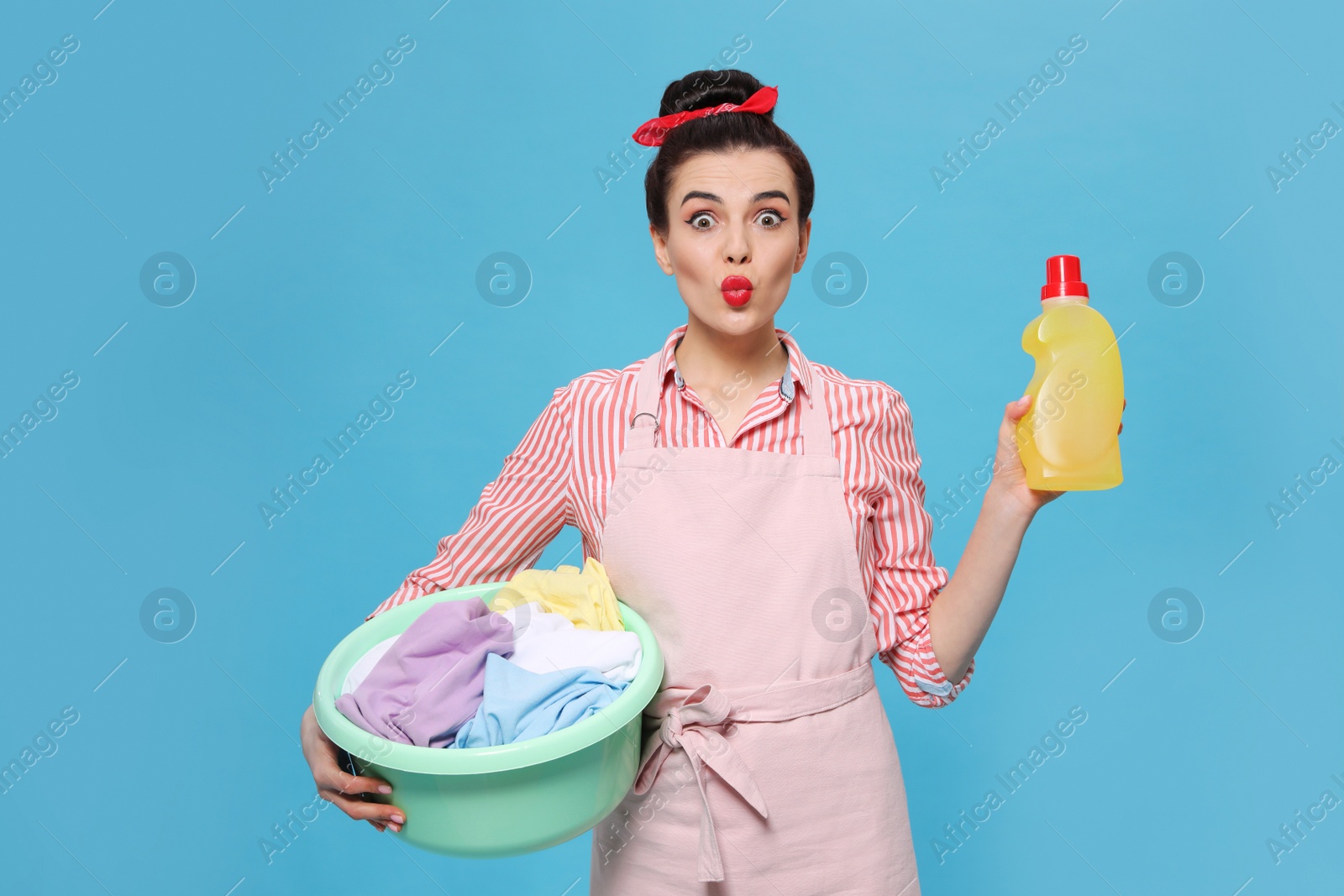 Photo of Housewife holding bottle of cleaning product and basin with clothes on light blue background