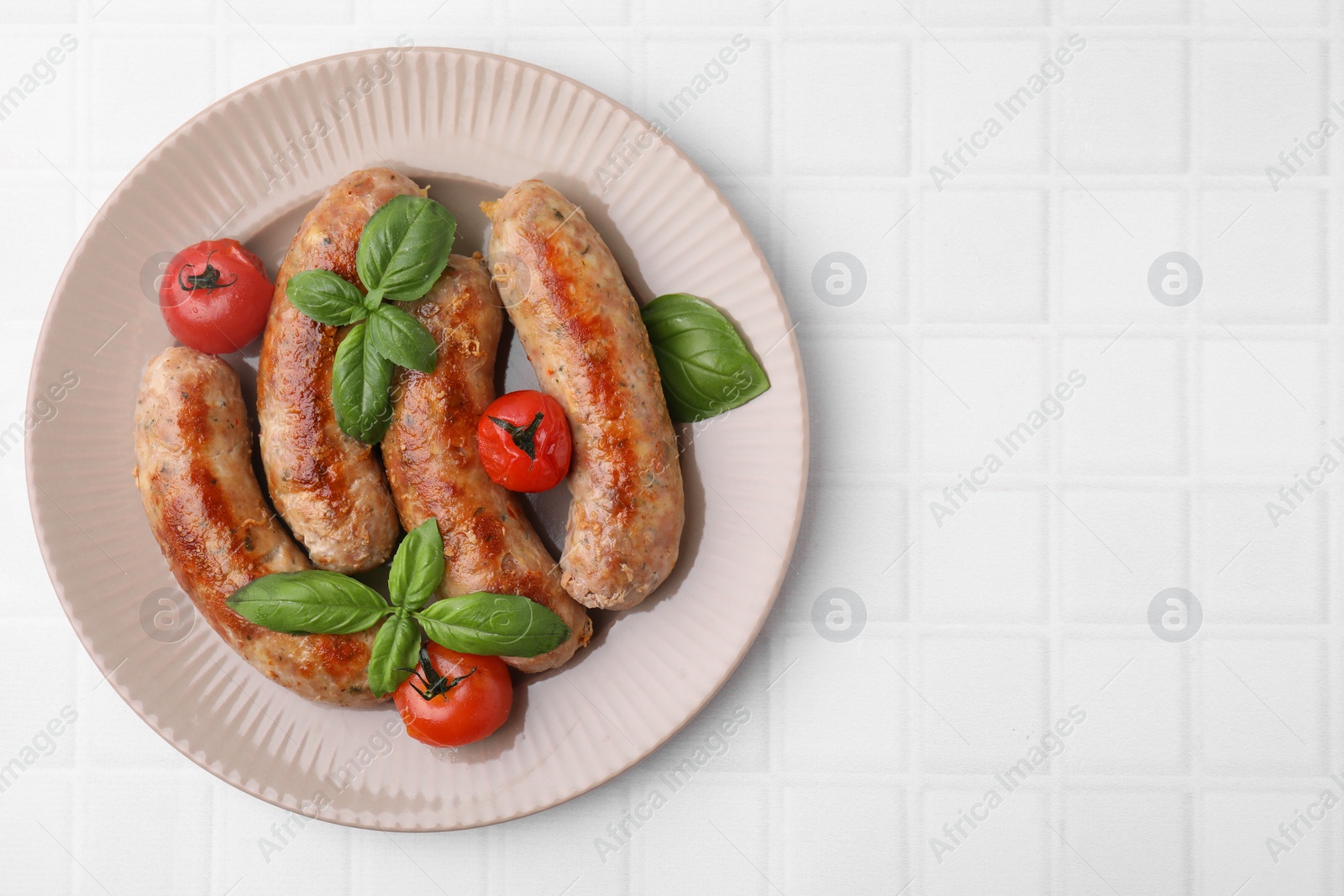 Photo of Plate with tasty homemade sausages, basil leaves and tomatoes on white tiled table, top view. Space for text