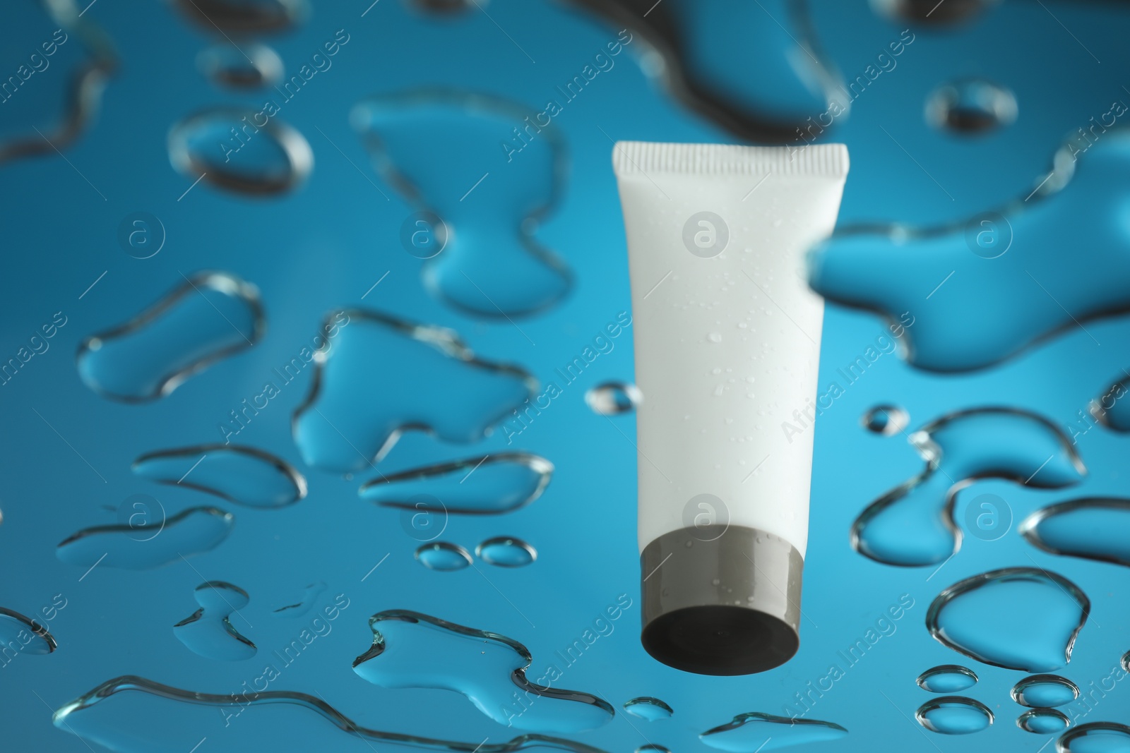 Photo of Moisturizing cream in tube on glass with water drops against blue background, low angle view. Space for text