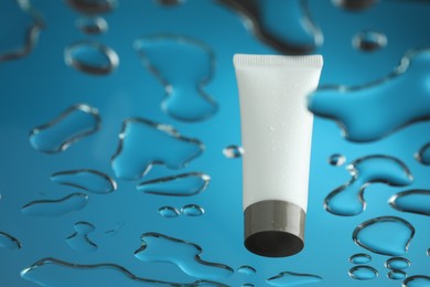 Moisturizing cream in tube on glass with water drops against blue background, low angle view. Space for text