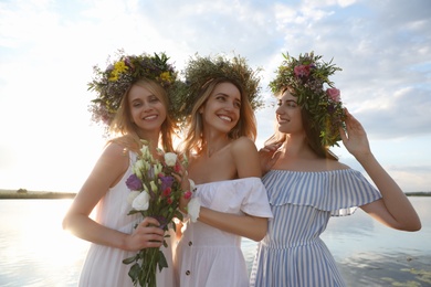 Young women wearing wreaths made of beautiful flowers near river on sunny day