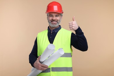 Photo of Architect in hard hat holding drafts and showing thumb up on beige background
