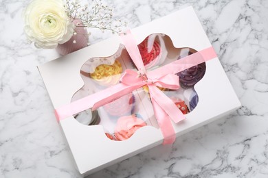 Photo of Different colorful cupcakes in box and flowers on white marble table, above view