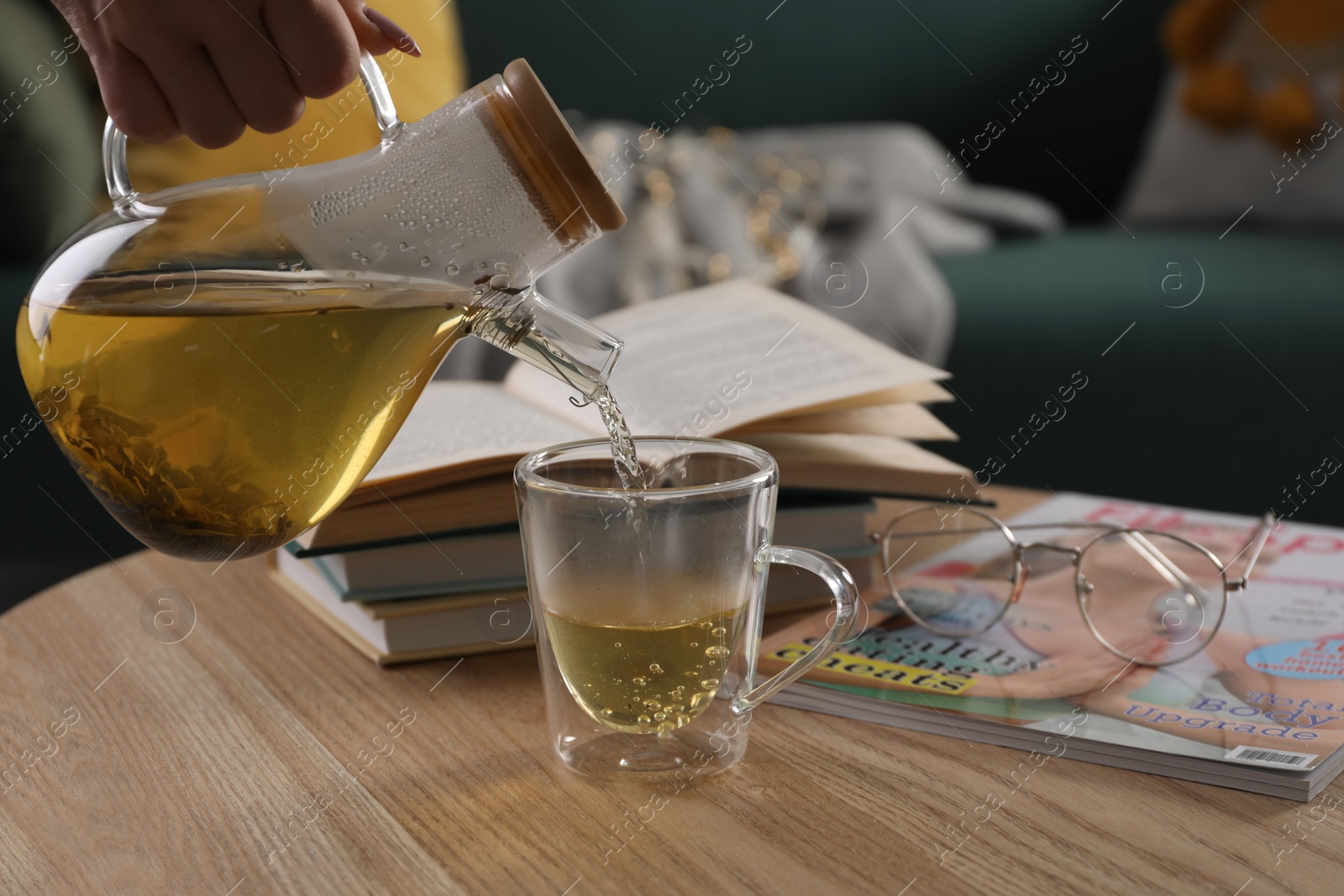 Photo of Woman pouring freshly brewed tea into cup at table in living room, closeup. Cozy home atmosphere