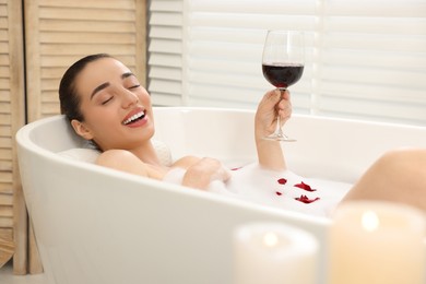 Happy woman with glass of wine taking bath in tub with foam and rose petals indoors