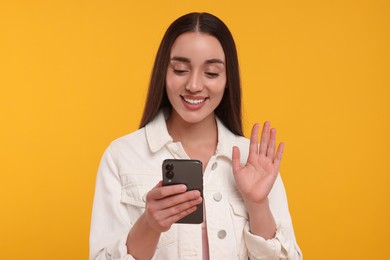 Happy young woman looking at smartphone and waving hello on yellow background
