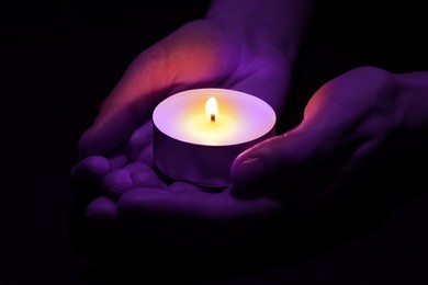 Woman holding burning violet candle in hands on black background, closeup. Funeral attributes