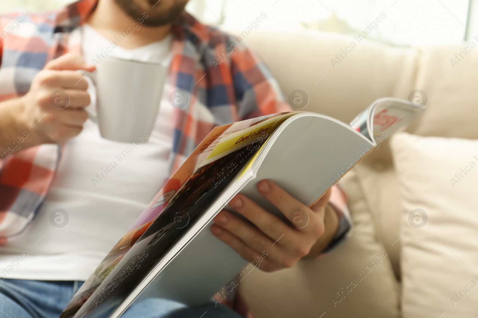 Photo of Man with cup of drink reading magazine on sofa, closeup