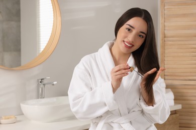 Photo of Happy young woman applying essential oil onto hair in bathroom