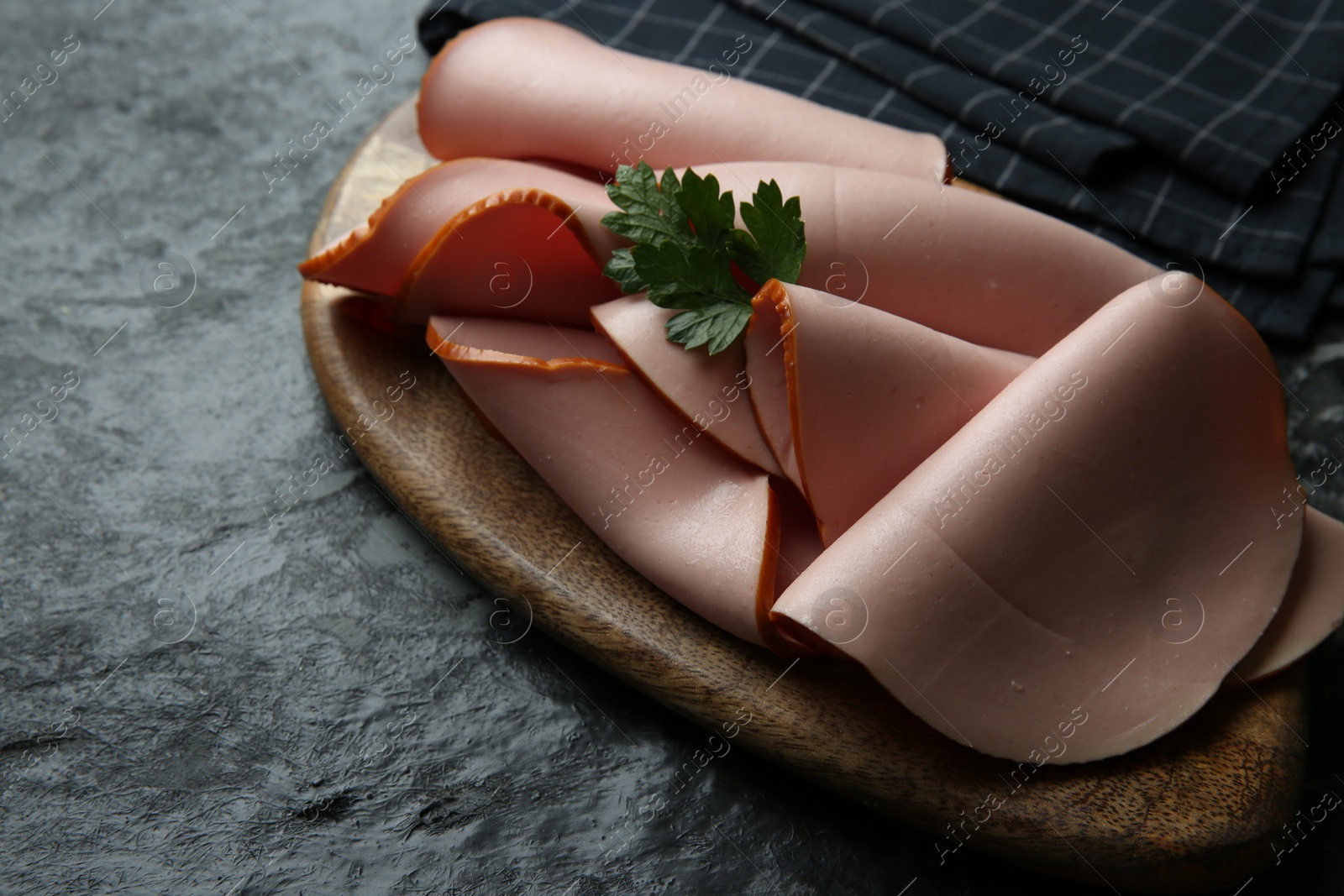 Photo of Board with slices of tasty boiled sausage and parsley on dark textured table, closeup