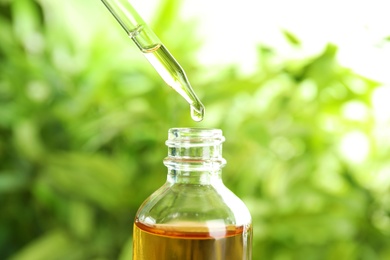 Photo of Essential oil dripping from pipette into glass bottle on blurred background, closeup