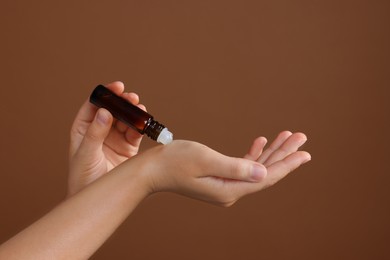 Photo of Woman with roller bottle applying essential oil onto wrist on brown background, closeup