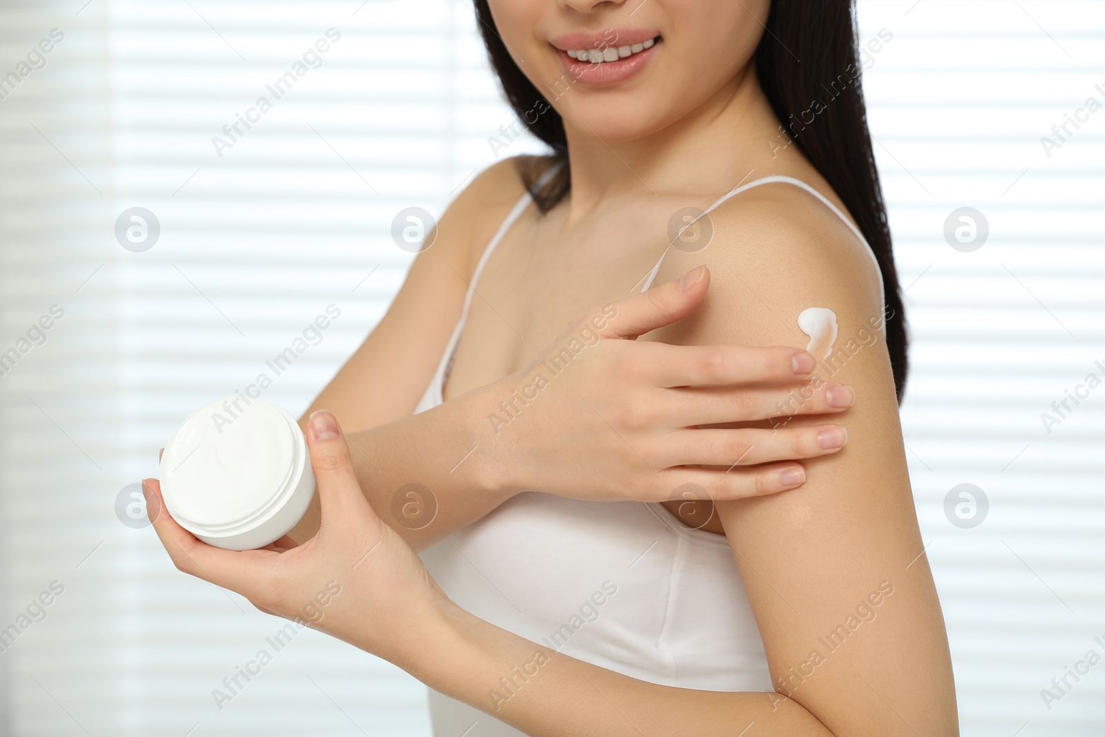 Photo of Young woman applying body cream on shoulder in room, closeup