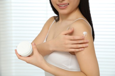 Photo of Young woman applying body cream on shoulder in room, closeup