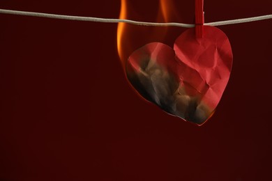 Crumpled red paper heart burning on rope against burgundy background, space for text. Broken heart
