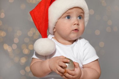 Cute baby in Santa hat with Christmas ball against blurred lights