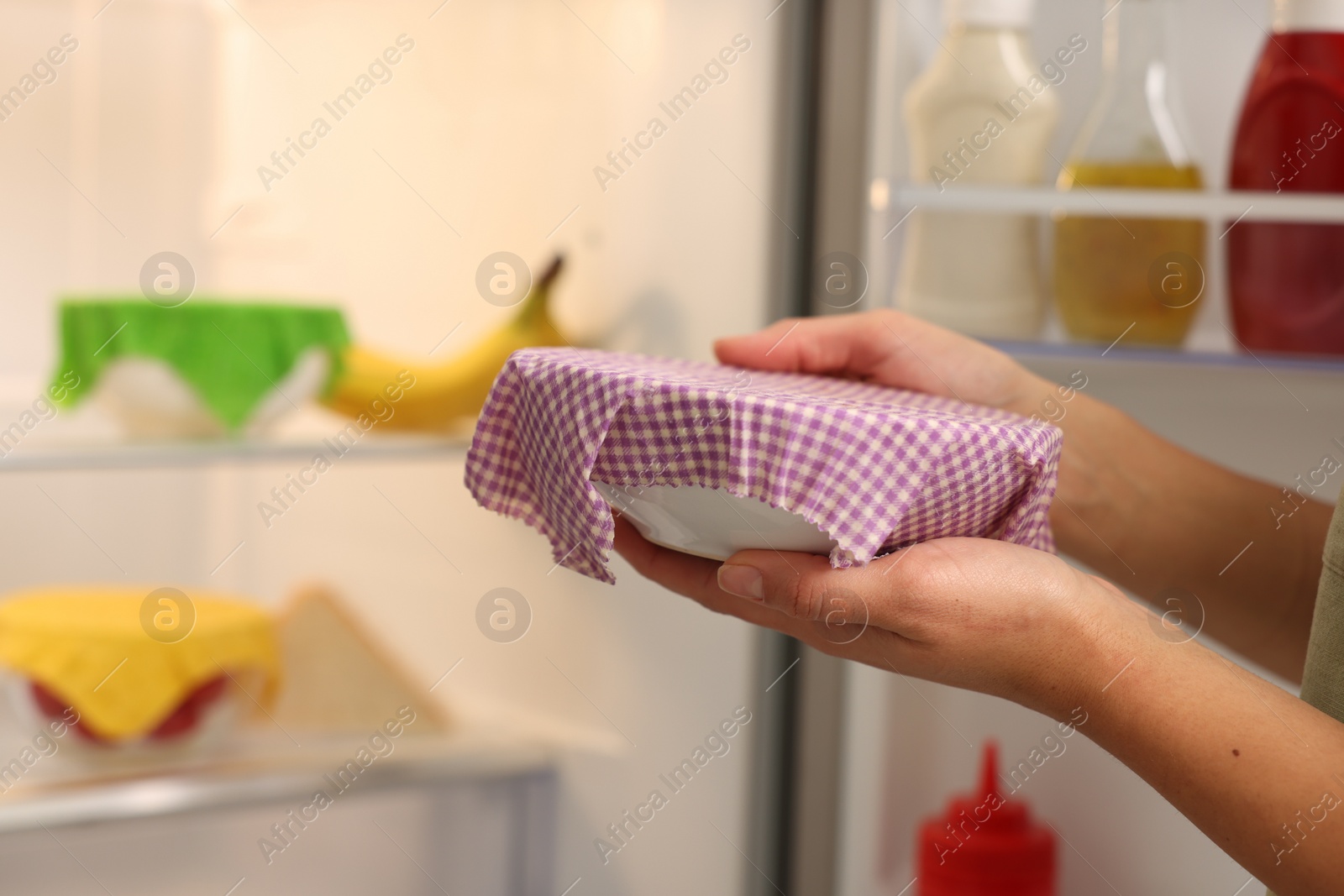 Photo of Woman holding bowl covered with beeswax food wrap near refrigerator in kitchen, closeup