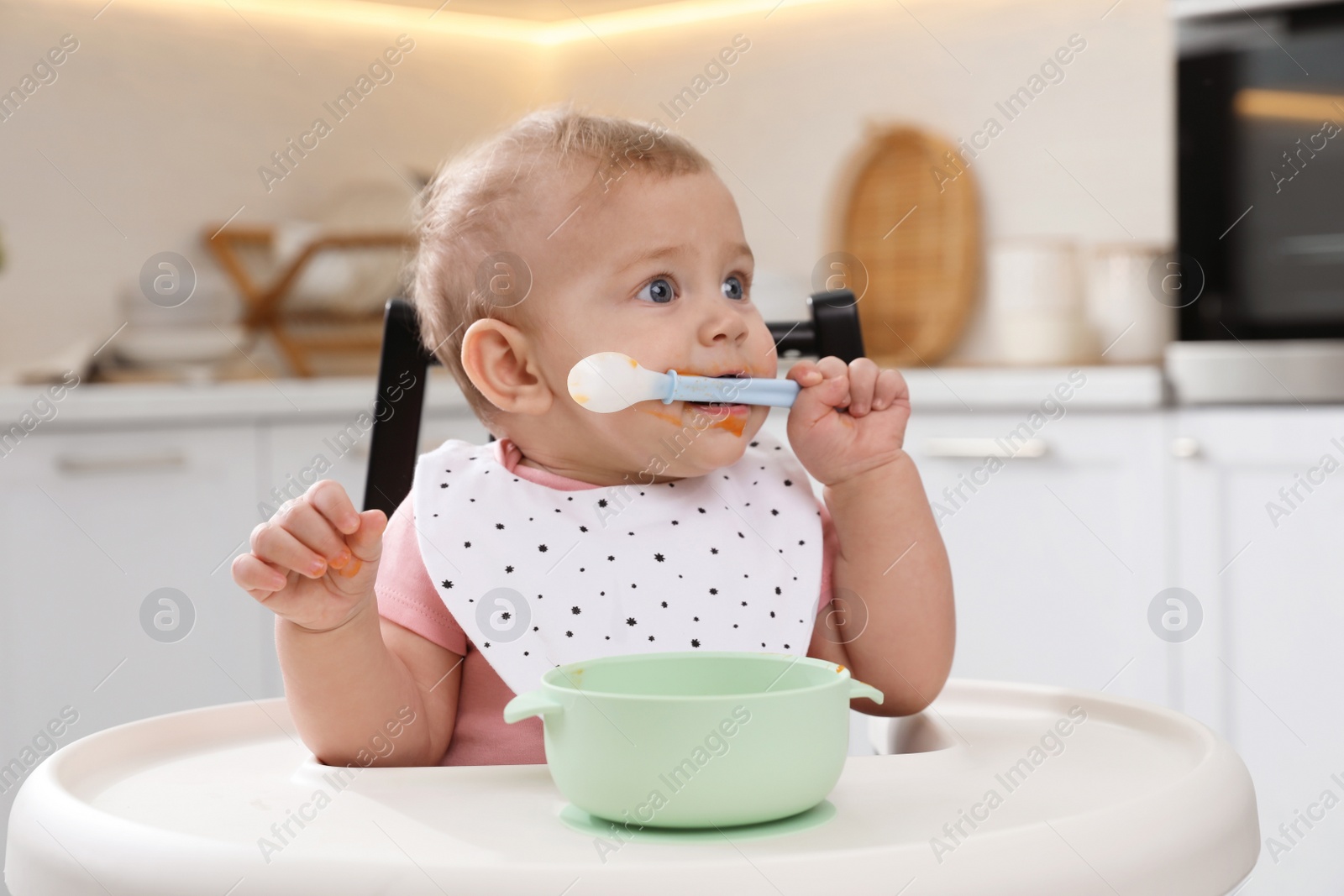 Photo of Cute little baby eating food in high chair at kitchen