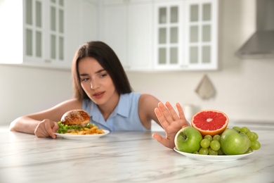 Photo of Woman choosing between fruits and burger with French fries in kitchen