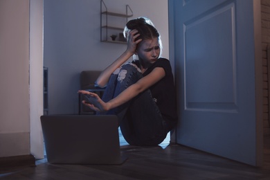 Photo of Frightened teenage girl with laptop on floor in dark room. Danger of internet