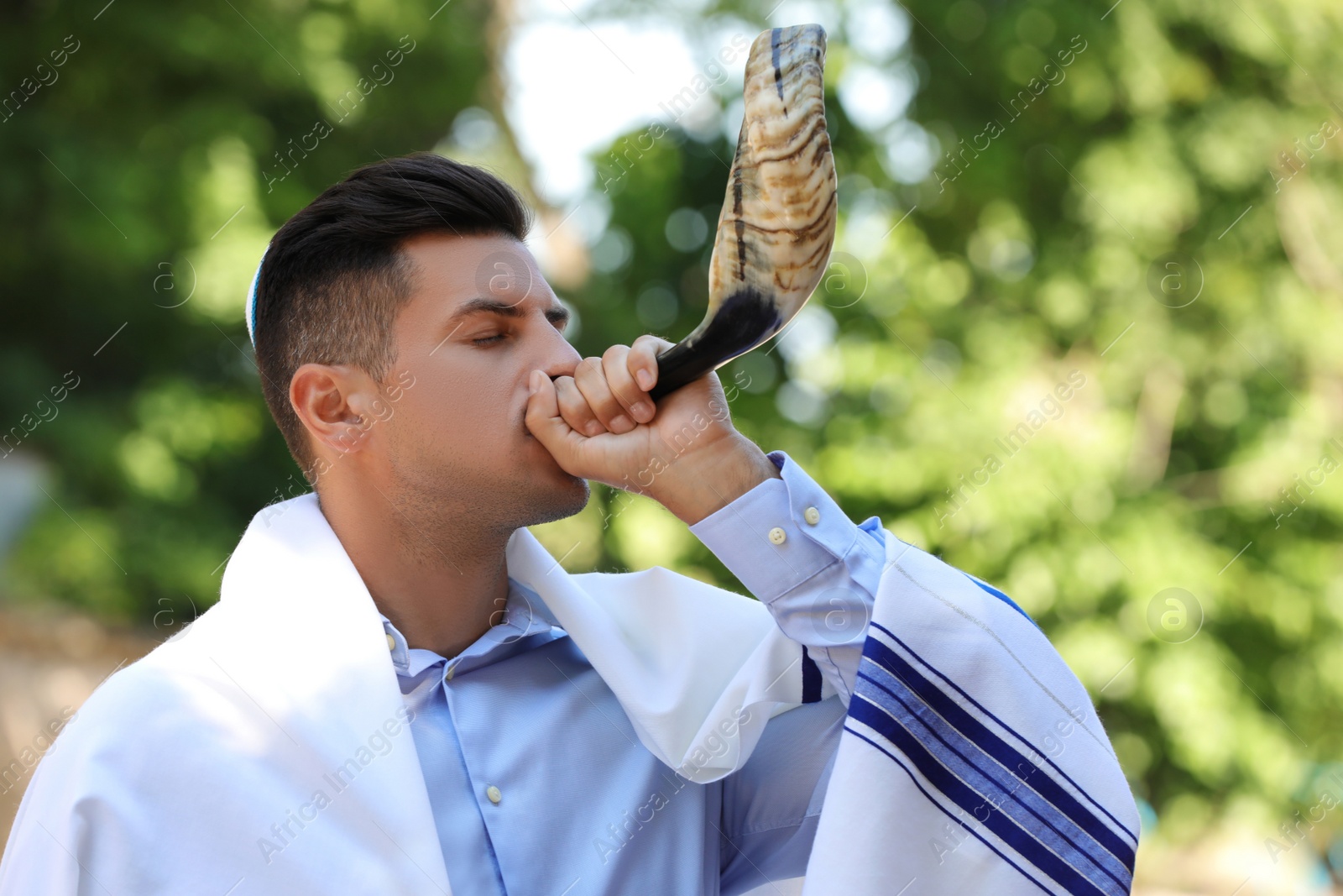 Photo of Jewish man in kippah and tallit blowing shofar outdoors. Rosh Hashanah celebration