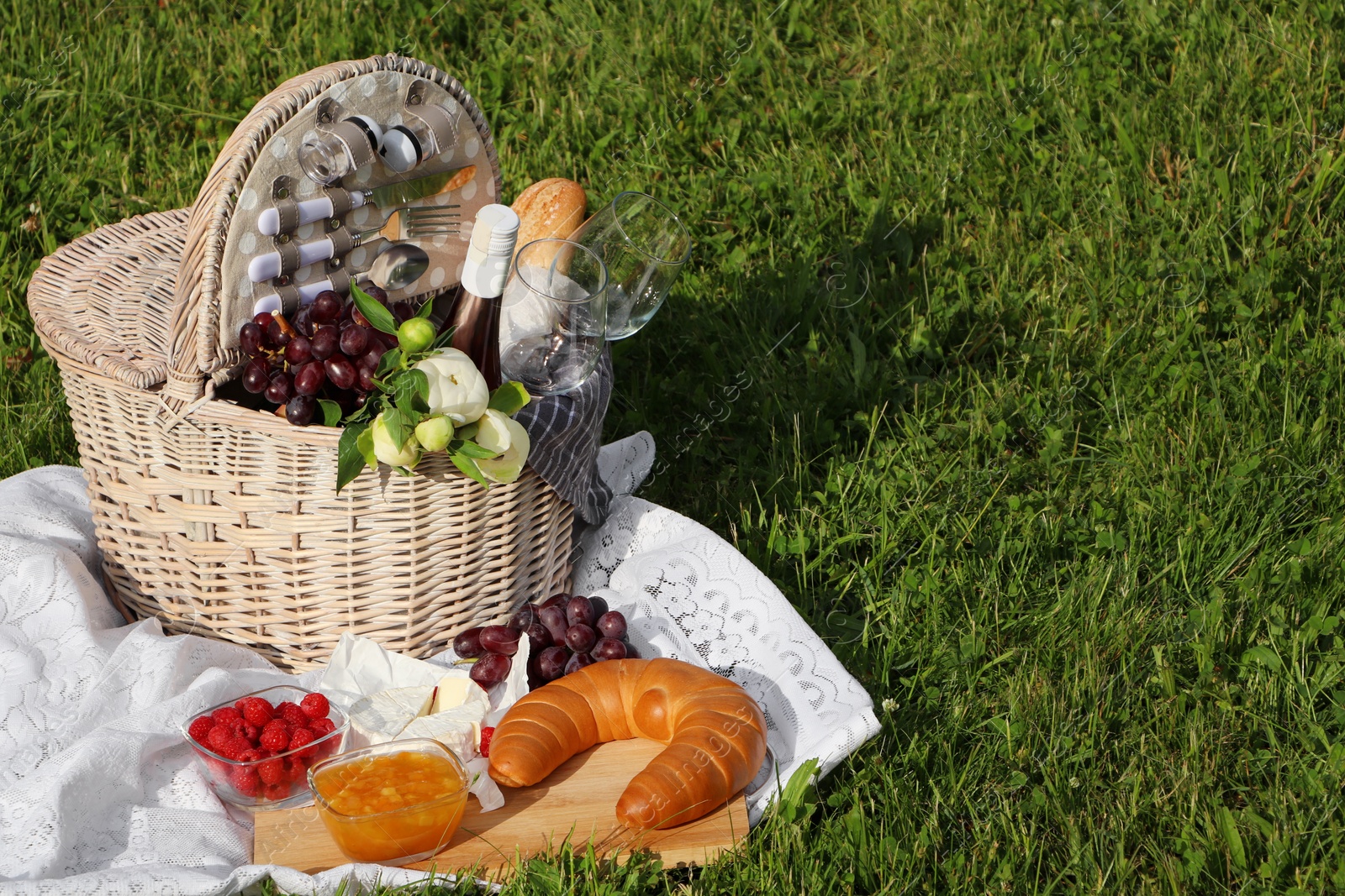 Photo of Picnic blanket with tasty food, flowers, basket and cider on green grass outdoors. Space for text