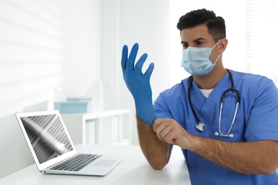 Photo of Doctor in protective mask putting on medical gloves at table in office