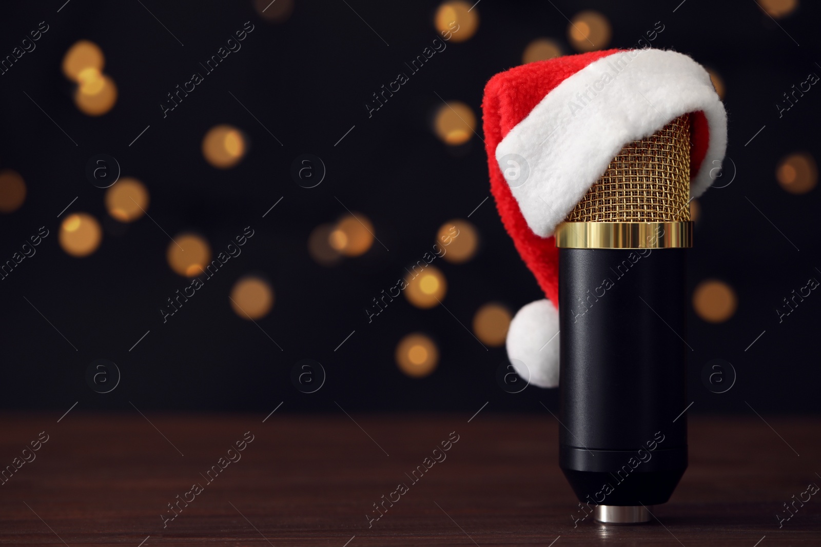 Photo of Microphone with Santa hat on wooden table against blurred lights, space for text. Christmas music