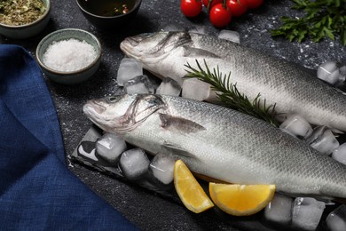 Photo of Fresh raw sea bass fish, ice cubes and lemon wedges on black table, closeup