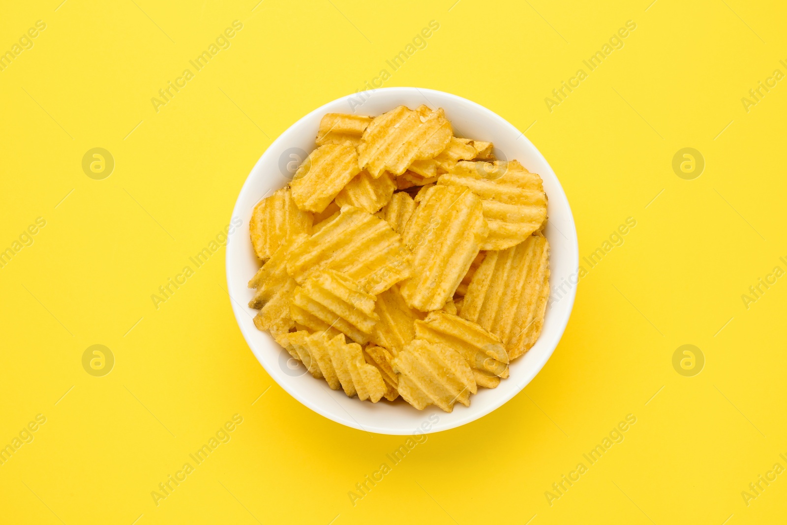Photo of Bowl of tasty ridged potato chips on yellow background, top view