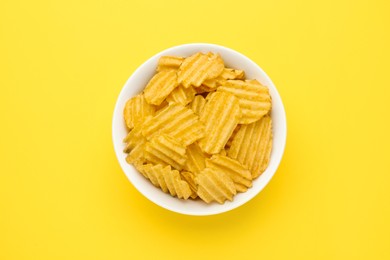 Bowl of tasty ridged potato chips on yellow background, top view