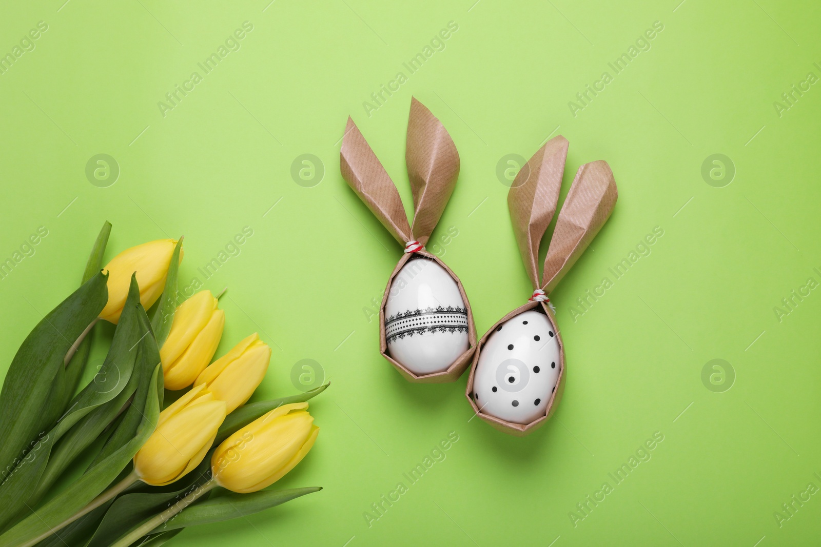 Photo of Easter bunnies made of craft paper and eggs near beautiful tulips on light green background, flat lay