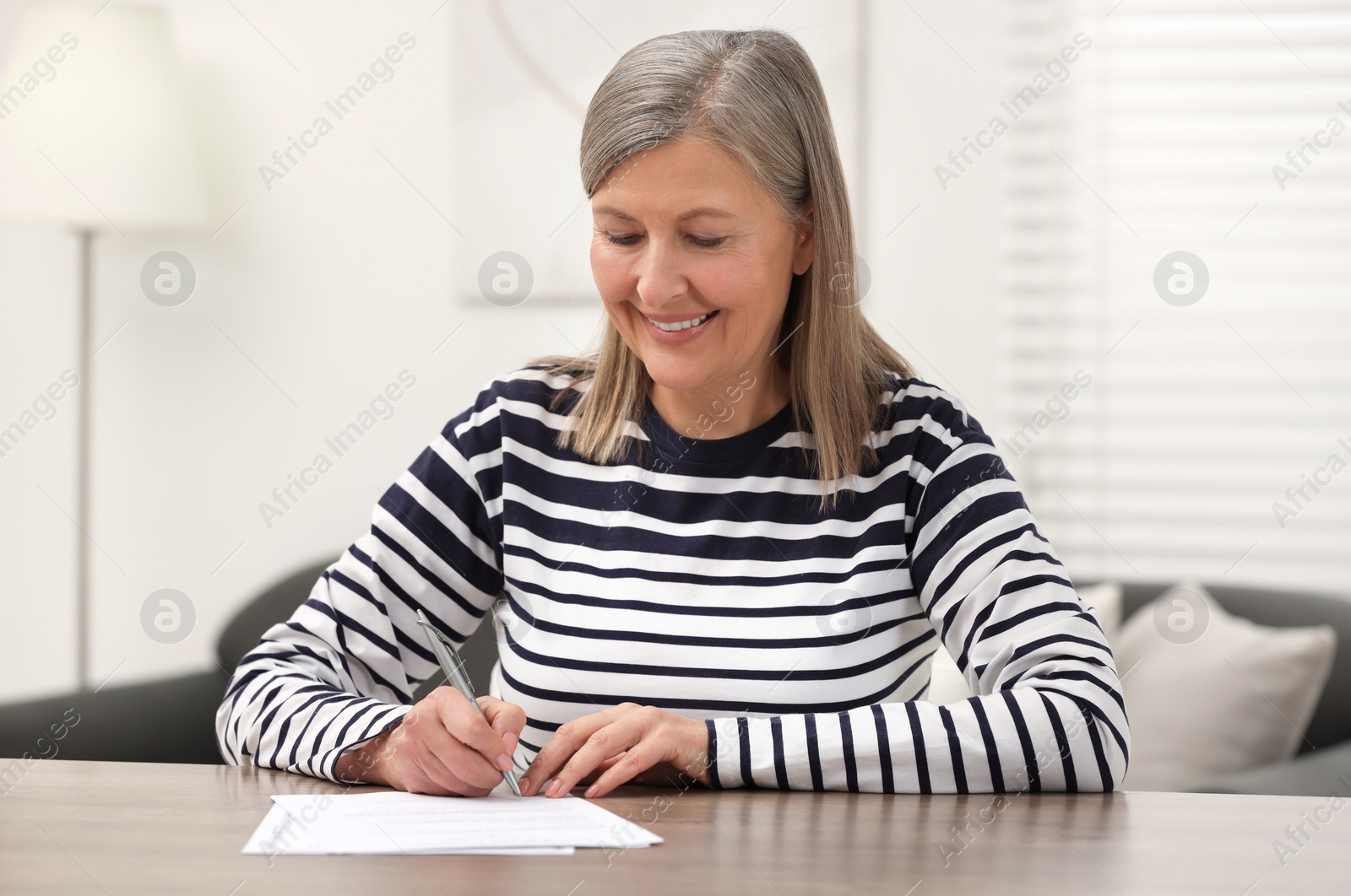 Photo of Smiling senior woman signing Last Will and Testament at wooden table indoors