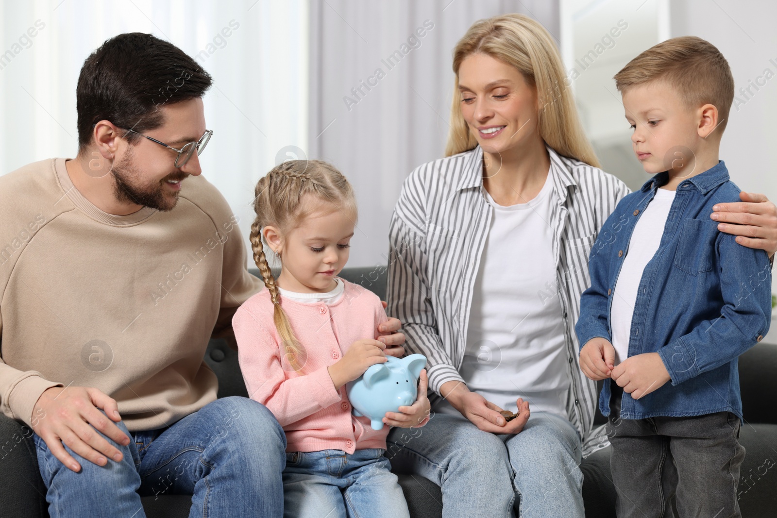 Photo of Family budget. Little girl putting coin into piggy bank while her parents and brother watching indoors