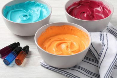 Photo of Bowls of different cream with food coloring and bottles of bright liquid on white wooden table