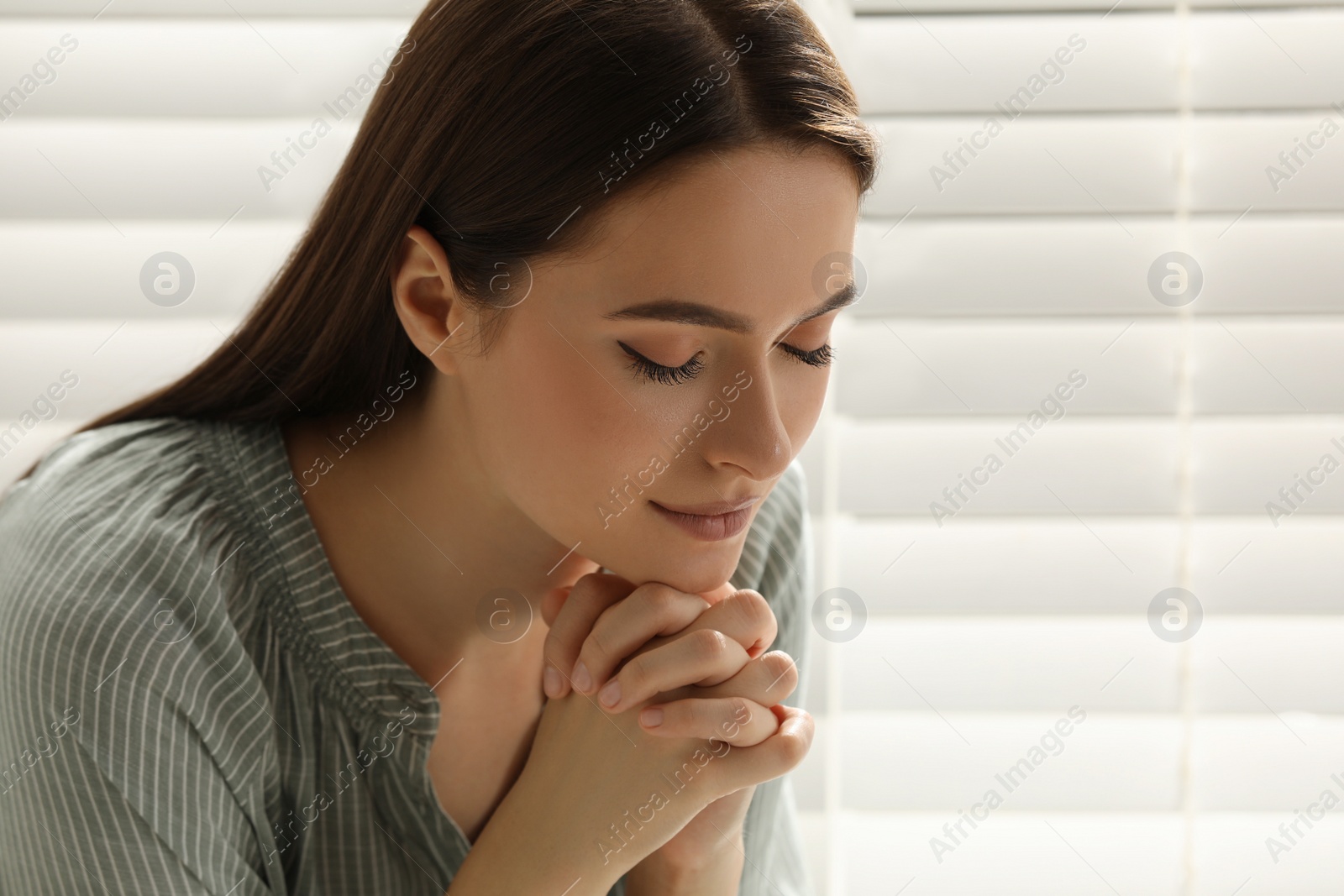 Photo of Religious young woman praying near window indoors, closeup