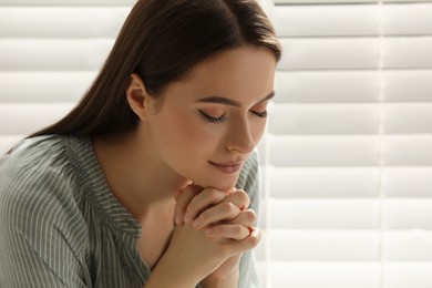 Religious young woman praying near window indoors, closeup