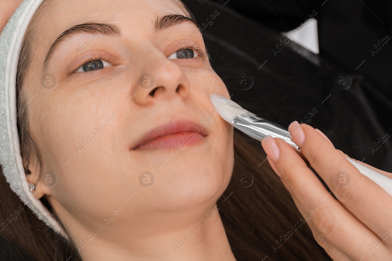 Photo of Cosmetologist applying mask on woman's face indoors, closeup