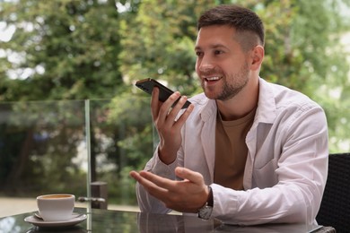 Handsome man recording voice message via smartphone at table in outdoor cafe. Space for text