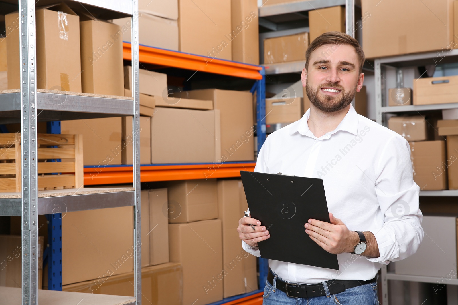 Photo of Young businessman with clipboard near rack of cardboard boxes at warehouse