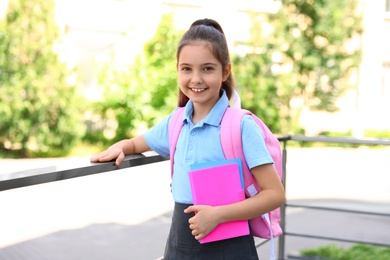 Cute little girl in school uniform with backpack and stationery near handrail outdoors