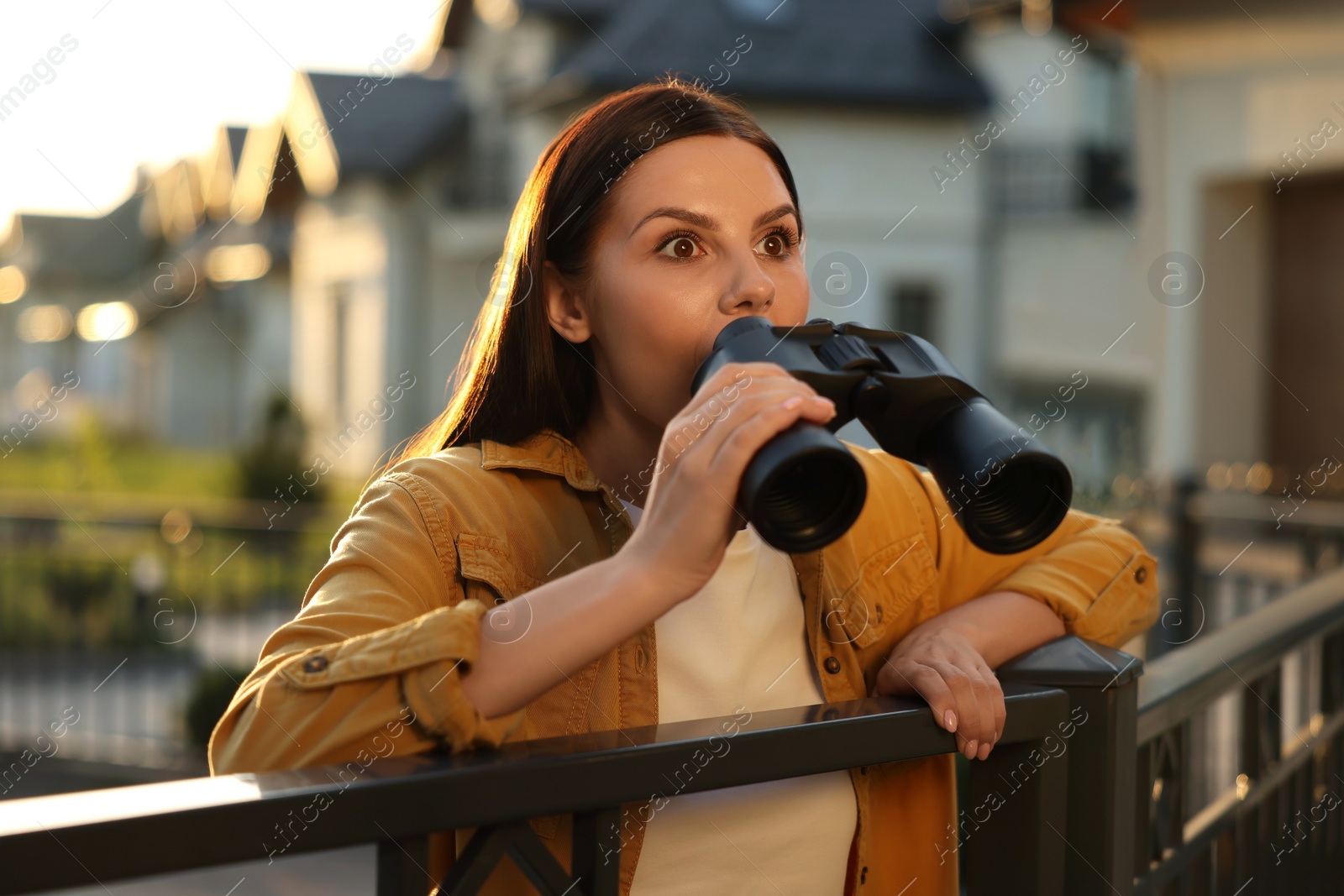 Photo of Concept of private life. Curious young woman with binoculars spying on neighbours over fence outdoors